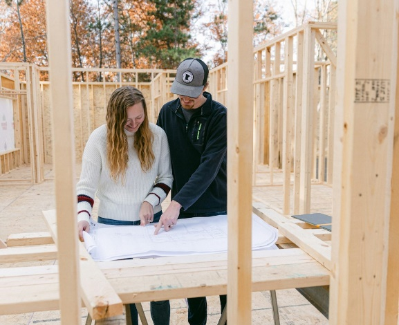 A couple reviewing construction plans on a table inside a wooden frame of a house under construction, highlighting the initial stages of building their Pennsylvania dream home.