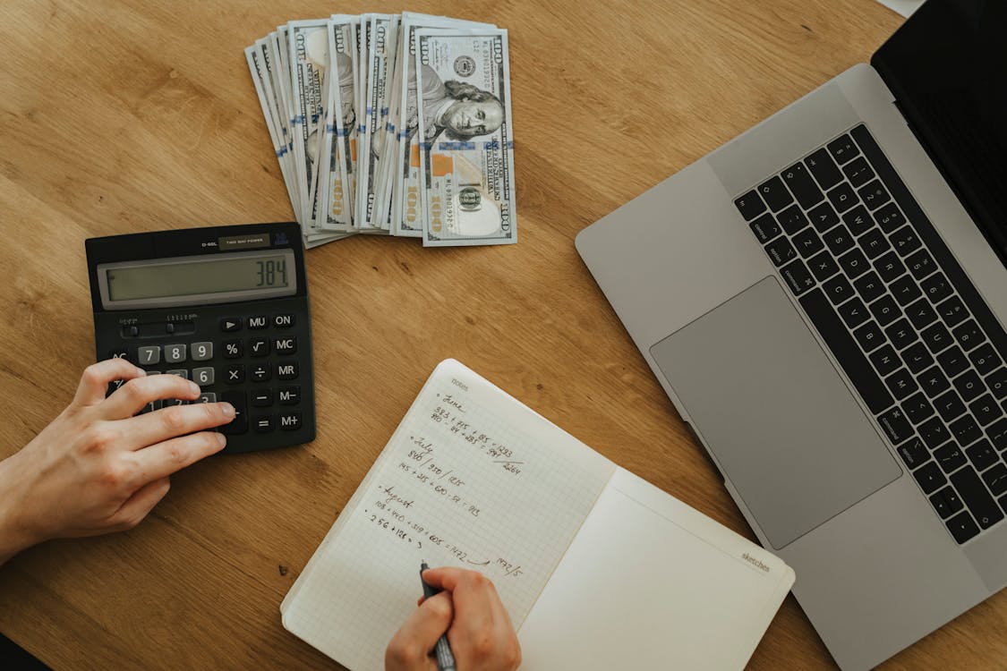 An image of a person using a calculator and writing in a notebook, with a bundle of 100 US dollar banknotes and a laptop on the table 