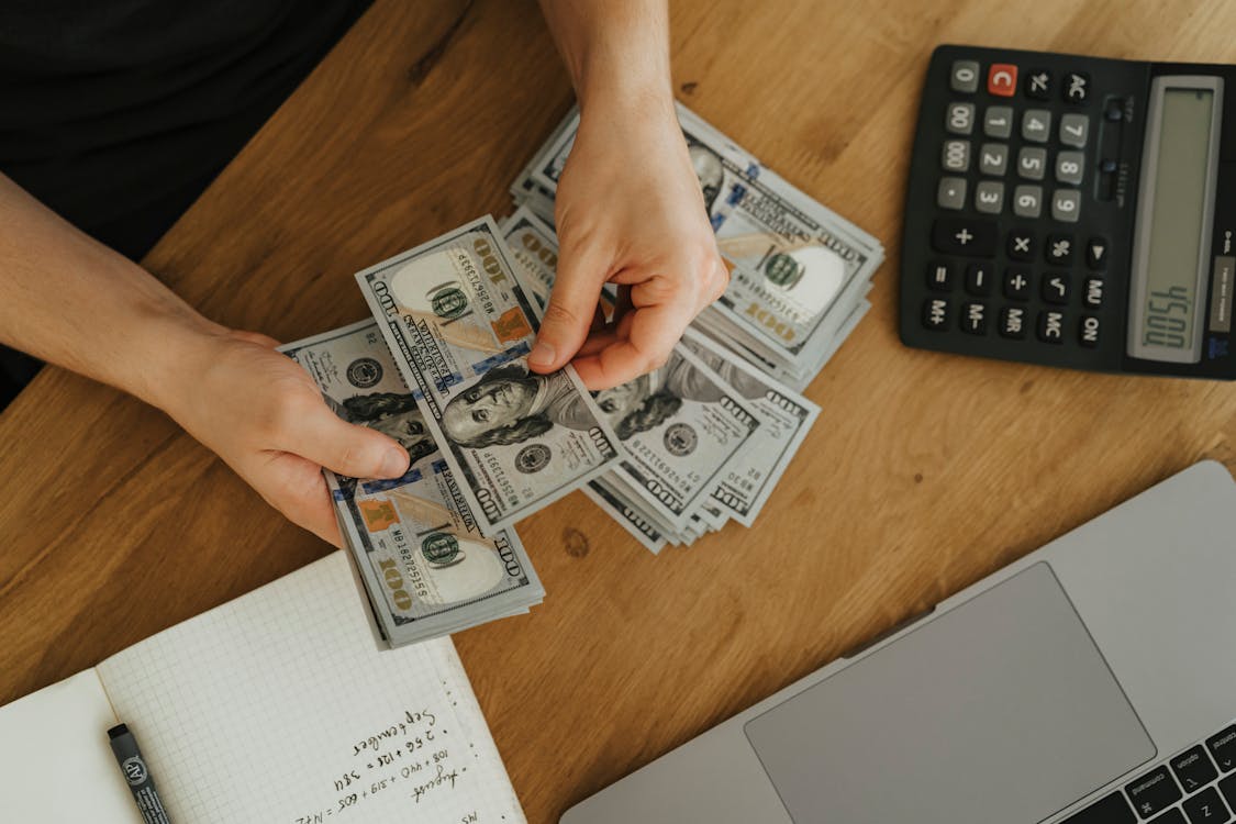 An image of a person counting 100 US dollar banknotes with a calculator on the desk