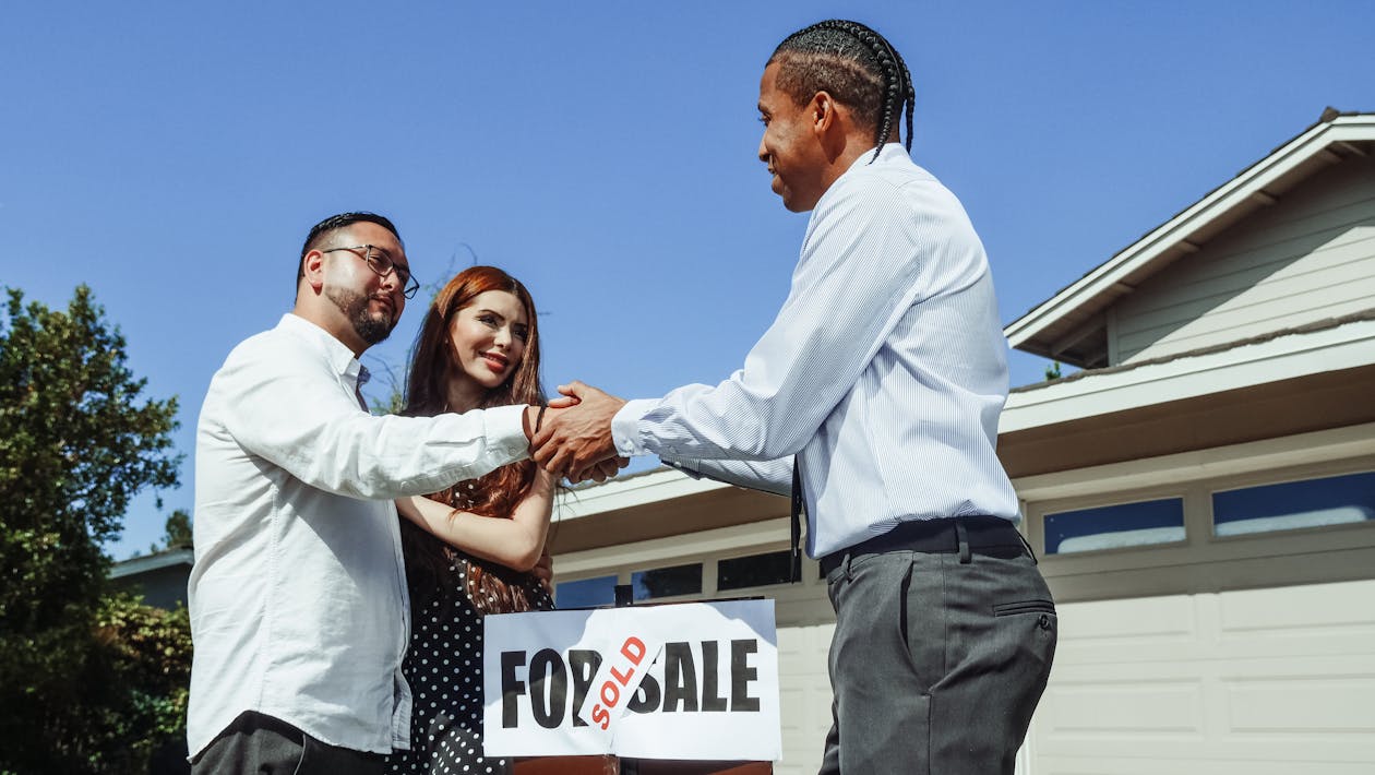 An image of a realtor shaking hands with his client above a sign that reads “Sold” on top of “For Sale”