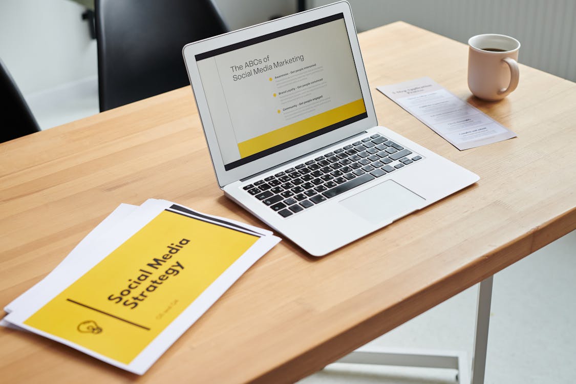 A close-up shot of a laptop beside paper documents with “Social media strategy” written on it