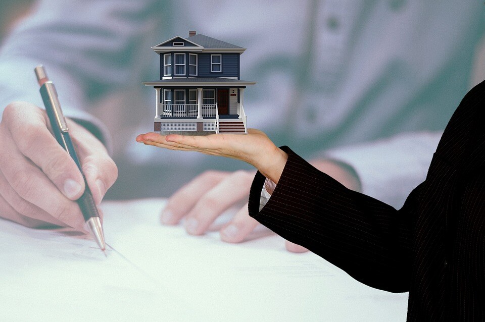 Hand holding a miniature house model, person signing paperwork.