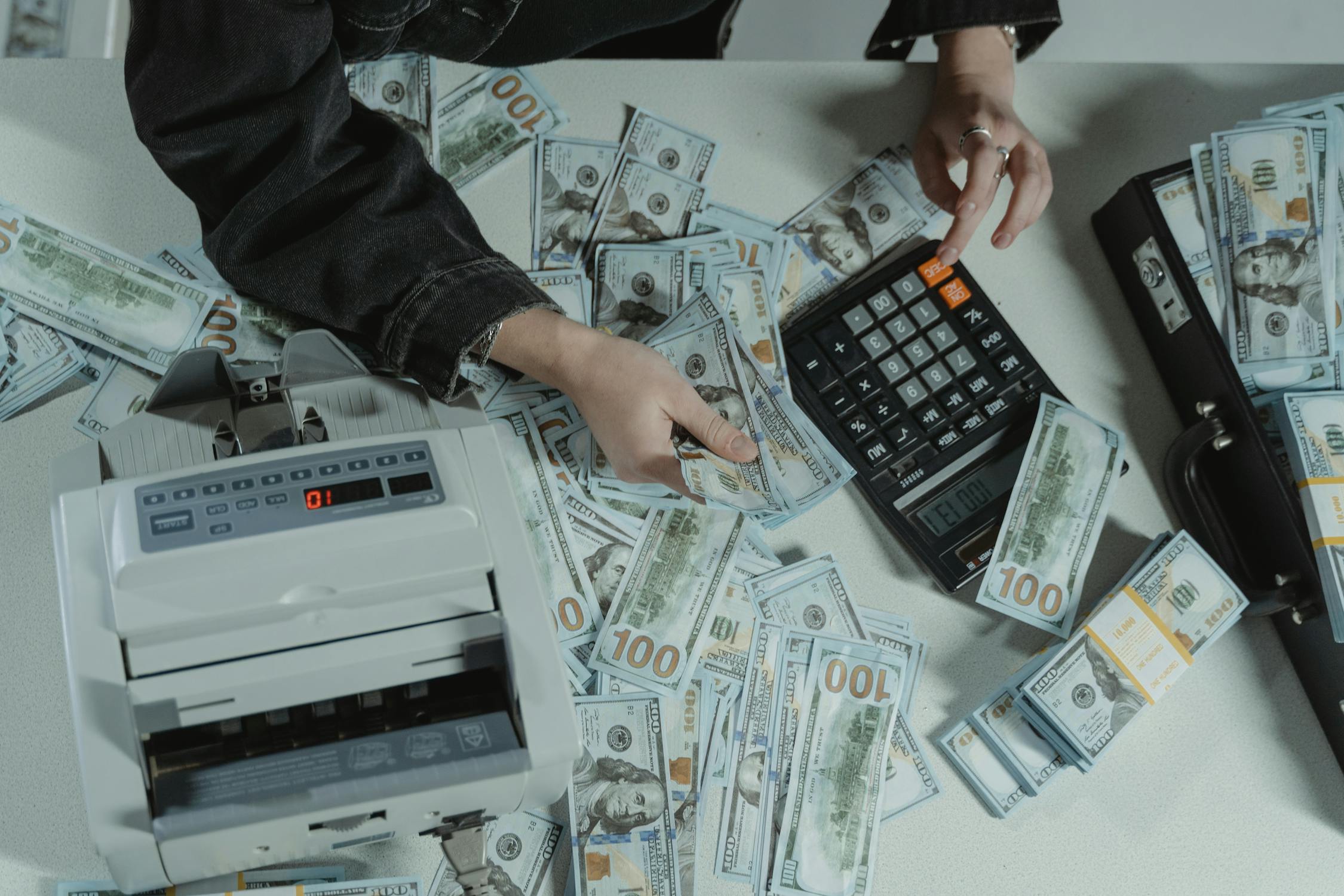 A person holding dollar bills while using a calculator with a money-counter machine on the table