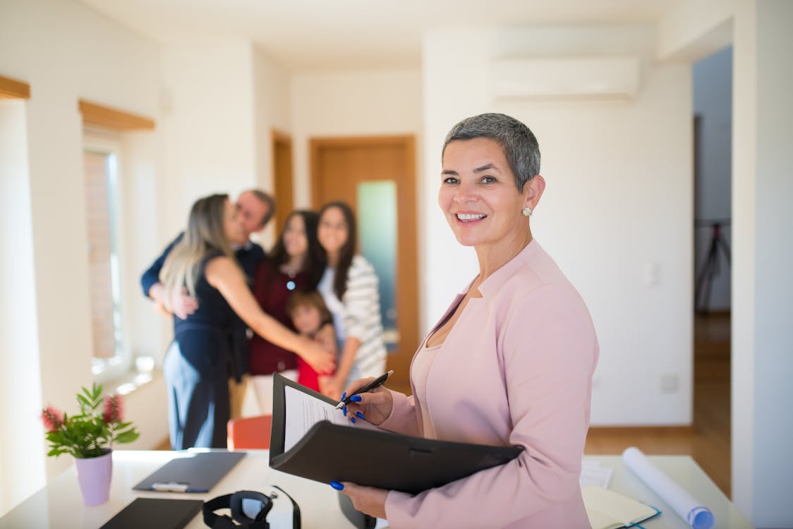 Real estate agent with clipboard smiling, family hugging in the background.